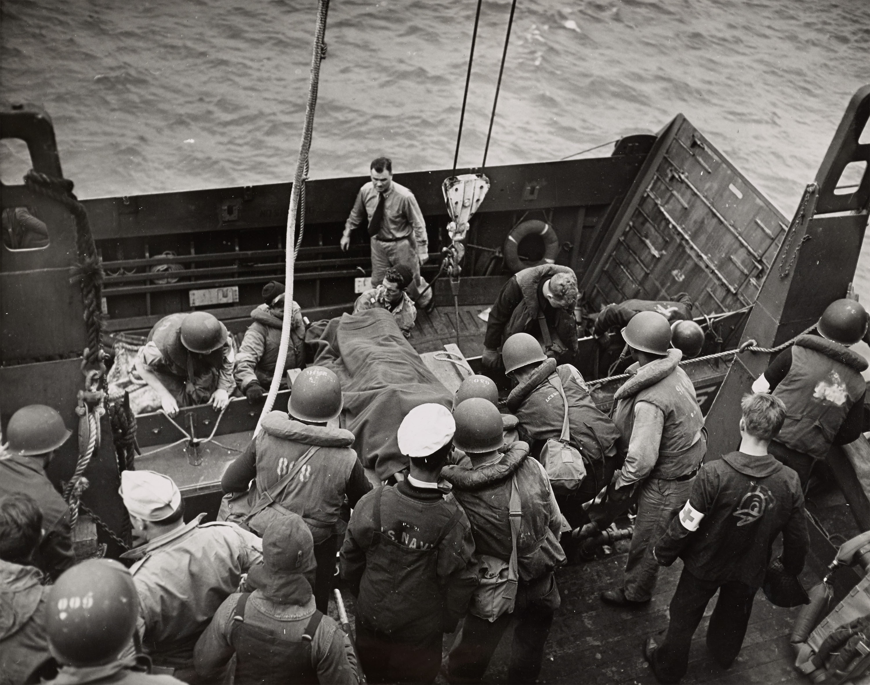 A group of Coast Guard gather around a stretcher on a ship’s deck, assisting a wounded person during a military operation at sea.